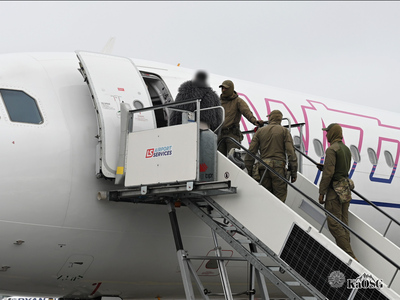 Na zdjęciu fragment samolotu na płycie lotniska. Kilku mężczyzn stoi na schodkach do samolotu. Ubrani są w mundury, twarze zasłonięte kominiarkami. Prawdopodobnie wyprowadzają z samolotu kobietę ubraną w szare futerko.