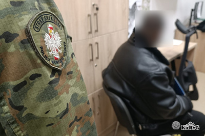 Na zdjęciu mężczyzna siedzący w pomieszczeniu biurowym. Ubrany w skórzaną kurtkę, Po jego lewej stronie widoczny fragment ramienia, mundur z plakietką z napisem Straż Graniczna.