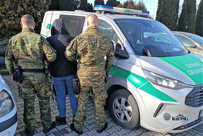 Dwóch funkcjonariuszy stoi tyłem z zatrzymanym na tle samochodu służbowego Straży Granicznej