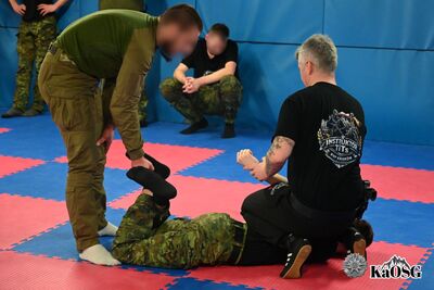 Instruktor na macie pokazuje sposób zabezpieczenia nóg i rąk osoby obezwładnionej. Funkcjonariusze w mundurach polowych przyglądają się precyzyjnym ruchom dłoni instruktora.