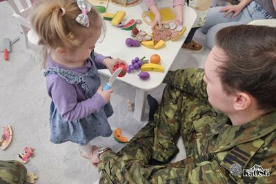 Na zdjęciu po prawej dolnej części strony funkcjonariusz ubrany w mundur moro, prawdopodobnie w pozycji siedzącej. Obok niego małe dziecko. W tle pomieszczenie prawdopodobnie przedszkole lub inna placówka dziecięca.