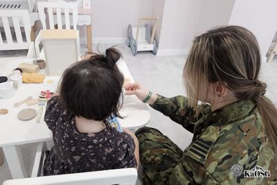 Na zdjęciu funkcjonariuszka ubrana w mundur moro. Obok niej małe dziecko. W tle widoczne zabawki i wyposażenie charakterystyczne dla placówki dziecięcej typu przedszkole.