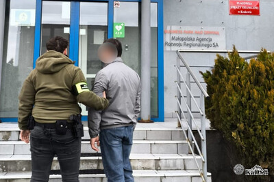 Na zdjęciu dwóch mężczyzn. Jeden - stojący z lewej strony- ubrany w czarne jeansy oraz bluzę z opaską na ramieniu z napisem straż graniczna. Mężczyzna posiada broń przy pasku i prowadzi pod rękę innego mężczyznę, prawdopodobnie osobę zatrzymaną.