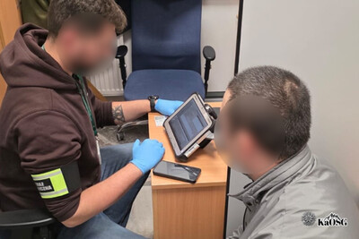 Na zdjęciu po lewej stronie mężczyzna siedzący na krześle z opaską na ramieniu z napisem straż graniczna. Mężczyzna spogląda na urządzenie elektroniczne typu czytnik. Po prawej stronie głowa i ramię innego mężczyzny prawdopodobnie osoby zatrzymanej. W tle krzesło biurowe.