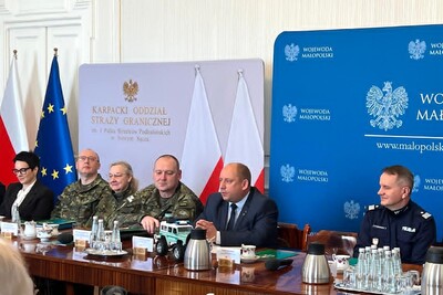 Grupa osób w mundurach i strojach cywilnych siedząca przy długim, drewnianym stole konferencyjnym. Na stole widać butelki wody, filiżanki oraz model terenowego samochodu Straży Granicznej. W tle flagi Polski i Unii Europejskiej.