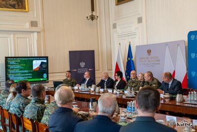 Szerokie ujęcie sali konferencyjnej w zabytkowym wnętrzu. Uczestnicy w mundurach siedzą tyłem do obiektywu, patrząc na ekran telewizora wyświetlający prezentację o aktach prawnych regulujących działanie Straży Granicznej.