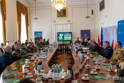Widok ogólny sali konferencyjnej z góry. Uczestnicy siedzą przy stole w kształcie litery U. Na środku ekran z prezentacją przedstawiającą statystyki i mapę. Nad stołem wisi kryształowy żyrandol.