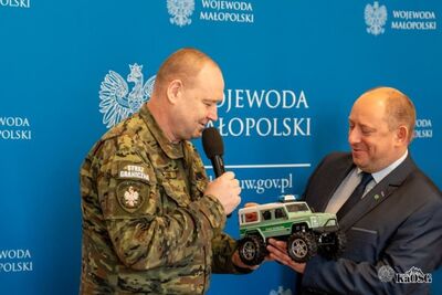 Funkcjonariusz Straży Granicznej trzyma mikrofon i wspólnie z mężczyzną w garniturze prezentuje model terenowego radiowozu Straży Granicznej. Tło stanowi niebieska ścianka z napisem „Wojewoda Małopolski”.