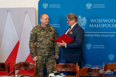 Mężczyzna w garniturze trzyma otwartą czerwoną teczkę i rozmawia z funkcjonariuszem Straży Granicznej w mundurze polowym. Stoją obok flagi Polski i niebieskiej ścianki promocyjnej.