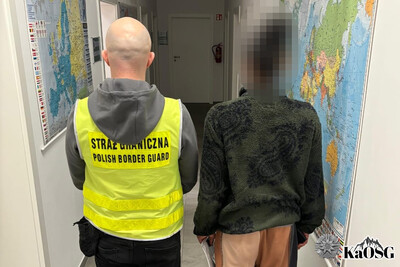 Funkcjonariusz w odblaskowej kamizelce z napisem straż graniczna polish border guard prowadzi zatrzymana cudzoziemkę korytarzem placówki sg w tarnowie. Obywatela USA trzyma w rękach wydaną decyzję o zobowiązaniu do powrotu oraz telefon komórkowy