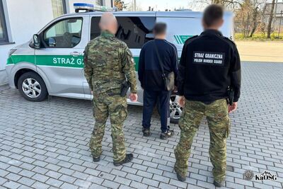 Na zdjęciu w środkowej części mężczyzna w granatowym dresie. Przez ramię ma przewieszoną męską, skórzaną torbę. Najprawdopodobniej jest to osoba zatrzymana. Obok niego, po obu stronach dwaj mężczyźni. Ten z lewej ma ubrany mundur moro, ten z prawej spodnie moro oraz czarny polar z napisem Straż Graniczna. W tle widoczne auto służbowe z kogutami i napisem Straż Graniczna.