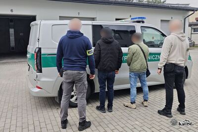 na zdjęciu czterech mężczyzn, dwóch z nich stojących po boku zdjęcia po prawej i lewej stronie, ma na ramionach opaski z napisem Straż Graniczna. Pomiędzy nimi dwaj inni mężczyźni, prawdopodobnie osoby zatrzymane. Wszyscy stoją przez samochodem służbowym typu więźniarka z bocznym napisem straż graniczna.