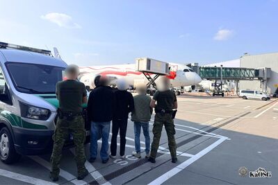 Na zdjęciu 5 mężczyzn, dwóch z nich po prawej i lewej stronie zdjęcia jest ubranych w spodnie moro i ciemnozielone koszulki z napisem na plecach straż graniczna. Pomiędzy nimi trzej inni mężczyźni, prawdopodobnie osoby zatrzymane. W tle płyta lotniska oraz samolot.