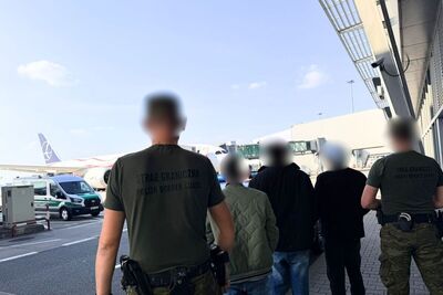 Na zdjęciu 5 mężczyzn, dwóch z nich po prawej i lewej stronie zdjęcia jest ubranych w spodnie moro i ciemnozielone koszulki z napisem na plecach straż graniczna. Pomiędzy nimi trzej inni mężczyźni, prawdopodobnie osoby zatrzymane. W tle płyta lotniska oraz samolot