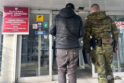 Na zdjęciu widać dwóch mężczyzn, którzy wchodzą do budynku urzędowego. Po lewej stronie widać czerwone tablice, które informują, że wchodzi się do budynku Placówki Straży Granicznej w Medyce.