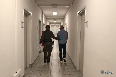 Na zdjęciu dwóch mężczyzn idących długim korytarzem. Mężczyzna po lewej stronie, ubrany w mundur i czapkę moro, w ręku trzyma dokumenty, prowadzi pod rękę innego mężczyznę ubranego w spodnie dżinsowej i koszulę oraz czapkę z daszkiem. W tle pomieszczenia biurowe.