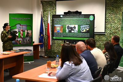 uczestnicy spotkania siedzą przy stołach, słuchają wystąpienia funkcjonariuszki, która na monitorze prezentuje dane dotyczące placówki sg w krakowie-balicach