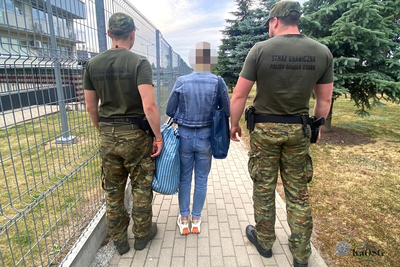 Na zdjęciu widać trzy osoby, które idą chodnikiem wzdłuż ogrodzenia. Dwie osoby ubrane są w mundury, a w środku idzie kobieta, która ma przez lewe ramię przewieszoną dużą torbę w niebieskim kolorze.