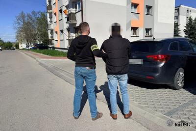 Dwie osoby stoją na parkingu tyłem. Przed nimi widać róg bloku mieszkalnego.