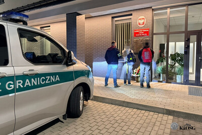 Na zdjęciu widać dwie osoby stojące przed budynkiem urzędu na którym widnieją czerwone tablice z białymi napisami. Po lewej stronie widać samochód oznakowany w kolorze srebrnym z zielonymi napisami i pasami.