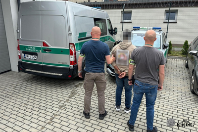 Na zdjęciu widać trzy osoby, które stoją tyłem. Mężczyzna w środku ma zamazaną głowę. Po lewej stronie widać samochód oznakowany w kolorze srebrnym z zielonymi napisami i pasami.