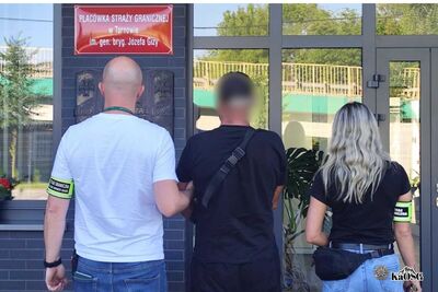 Po lewej stronie widoczny mężczyzna w ubraniu cywilnym z opaską na ramieniu z napisem straż graniczna, po prawej kobieta także ubrana po cywilnemu z opaską straż graniczna. Oboje prowadza mężczyznę - prawdopodobnie osobę zatrzymaną W tle budynek ze szklanymi drzwiami. Po jego lewej stronie godło Polski oraz metalowa tabliczka z napisem Placówka Straży Granicznej w Tarnowie.