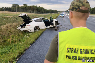 na pierwszym planie plecy funkcjonariusza w kamizelce odblaskowej, w tle rozbity biały samochód w rowie