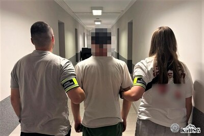 Funkcjonariuszka i funkcjonariusz w stroju cywilnym - z odblaskowymi opaskami na ramionach z napisem Straż Graniczna - prowadzą zatrzymanego cudzoziemca długim korytarzem